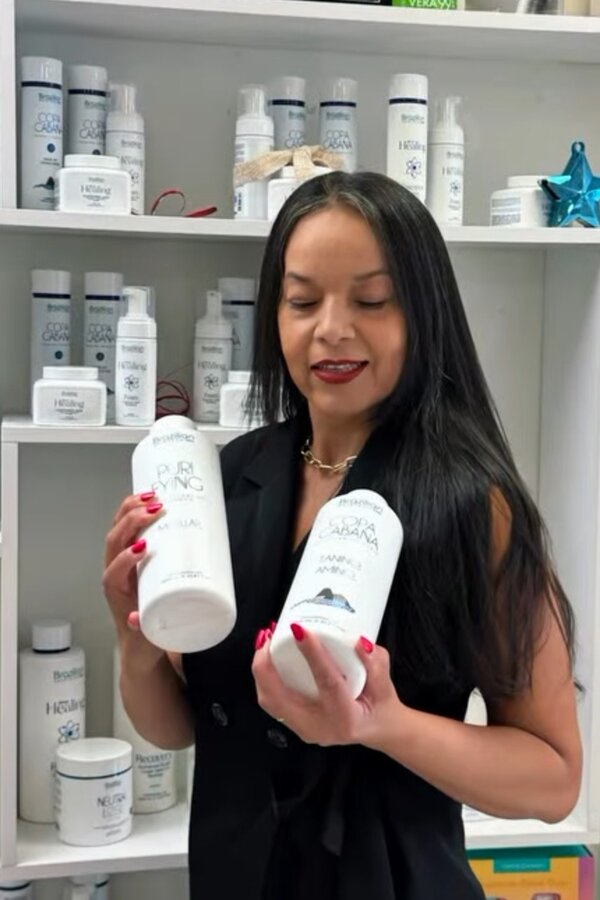 Eva Saturno in the salon holding Purifying and Copa Cabana Brazilian Protein bottles, with stocked product shelves behind her
